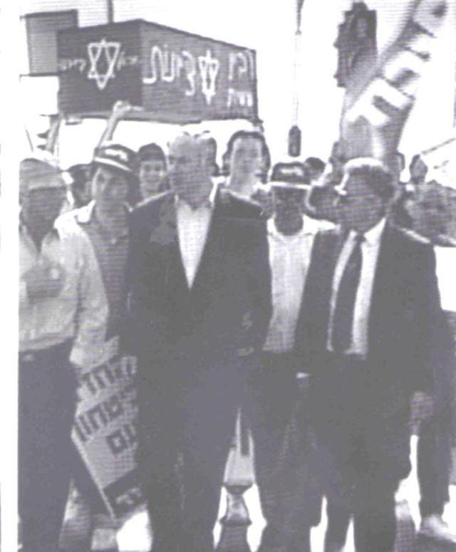 gray, blurry photograph of a crowd of people holding up a coffin on which stars of david and the words rabin and tzionut are written in hebrew, and the image of a younger than today bibi netanyahu in a suit jacket, looking ready to incite murder and derail the freedom of a whole people