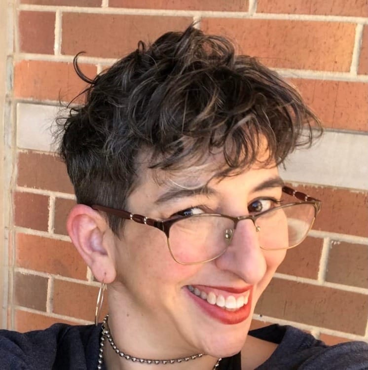 Photo of a white Jew with short dark hair with grey streaks, with glasses, red lipstick, a grin, silver hoop earirings