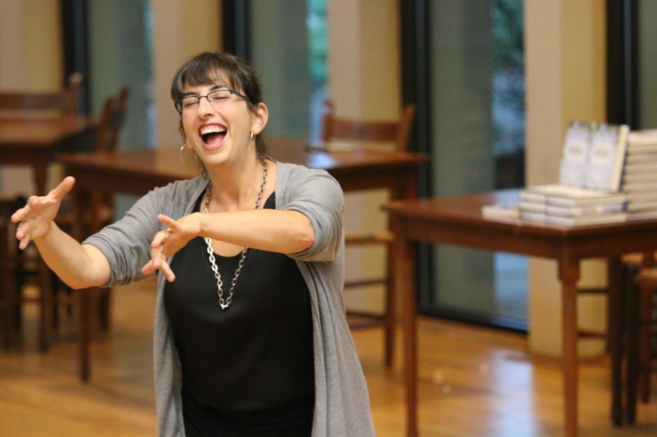 Danya teaching, brown bangs, grey cardigan, black tank top, chunky silver necklace, caught talking with her hands and a smile on her face