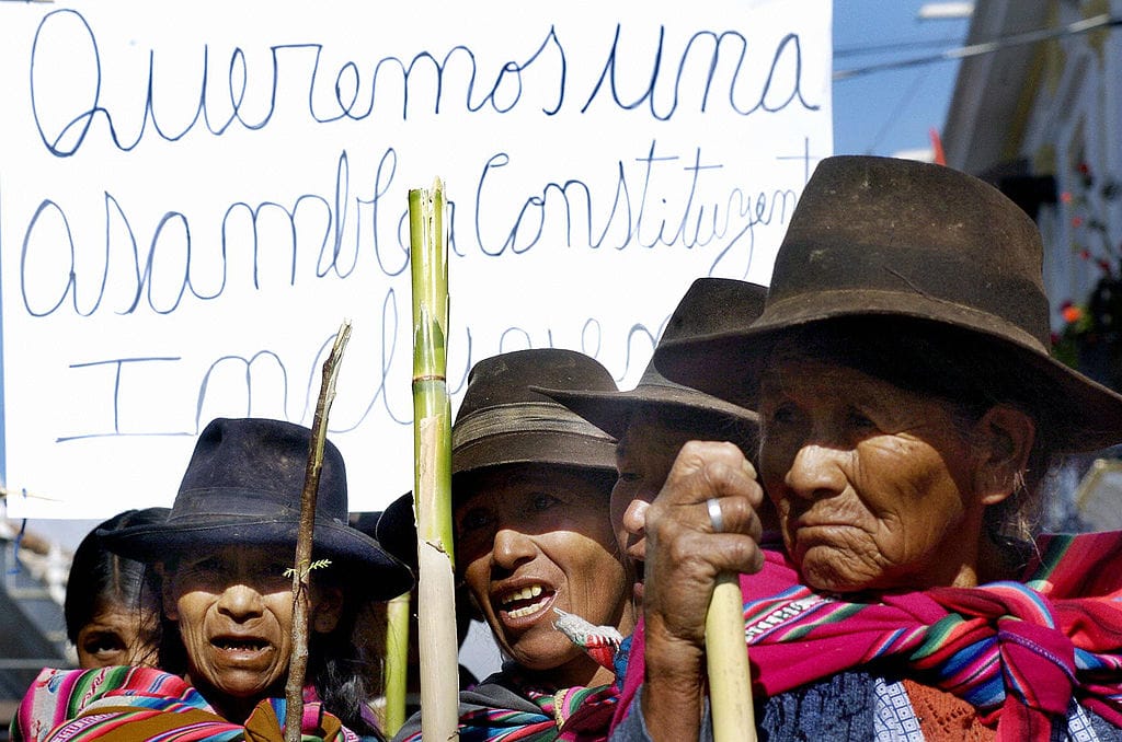 Quechua people holding a sign demanding a constituent assembly, wearing colorful woven shawls and brown and black hats.