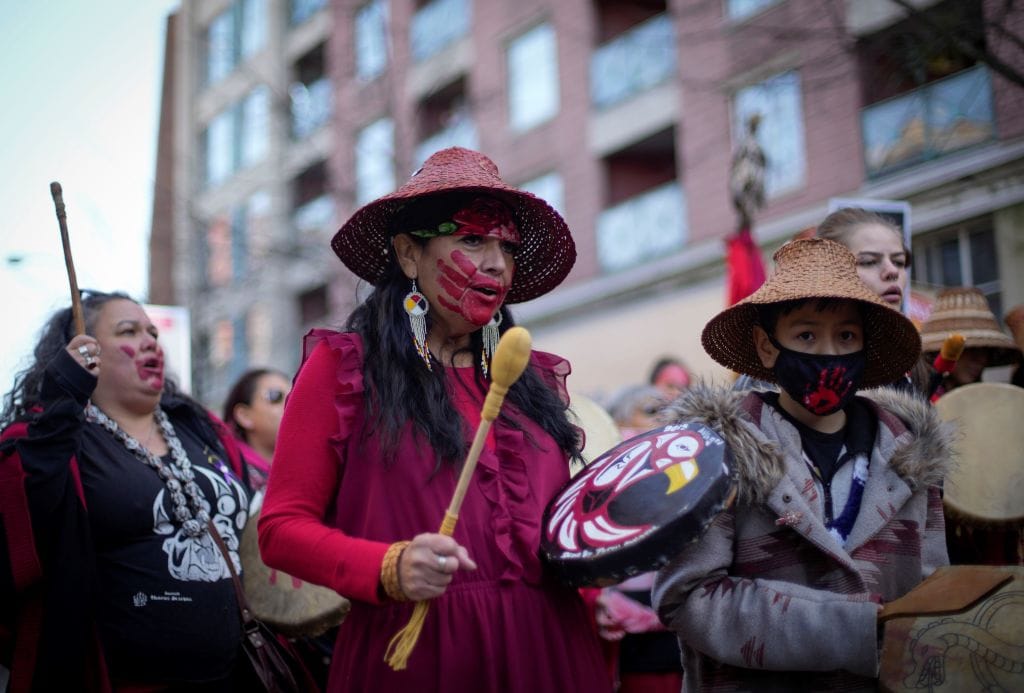 A number of First Nations people with drums and red hands painted over their faces