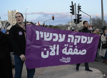 Guys hold up a purple banner in Hebrew and Arabic