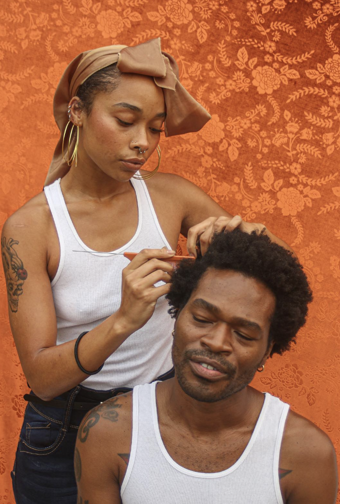 A Black woman with jeans and a tank top and hair in a scarf parts the head of a Black man, whose eyes are closed.